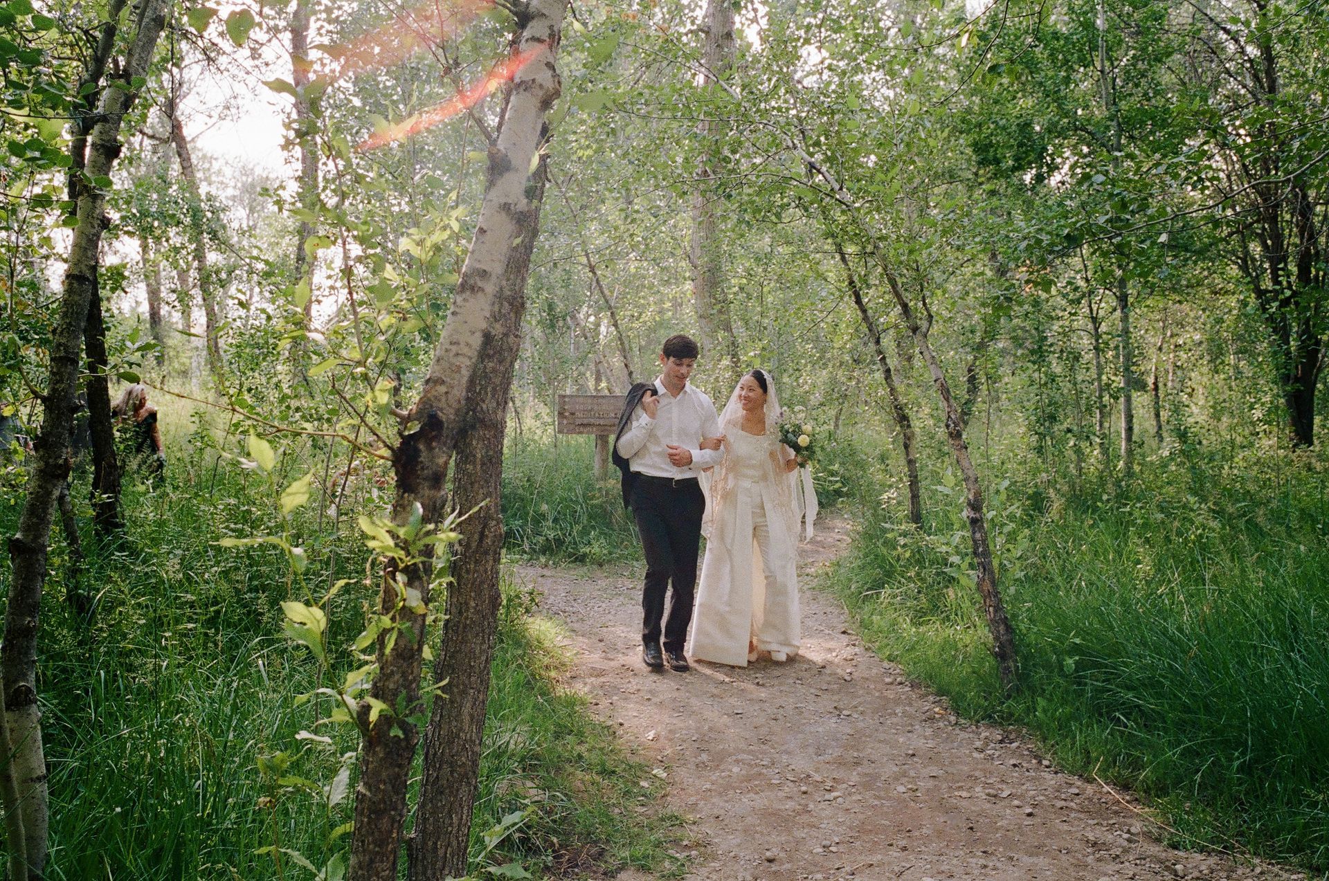 Two people walking arm-in-arm on a forest path, surrounded by greenery and sunlight