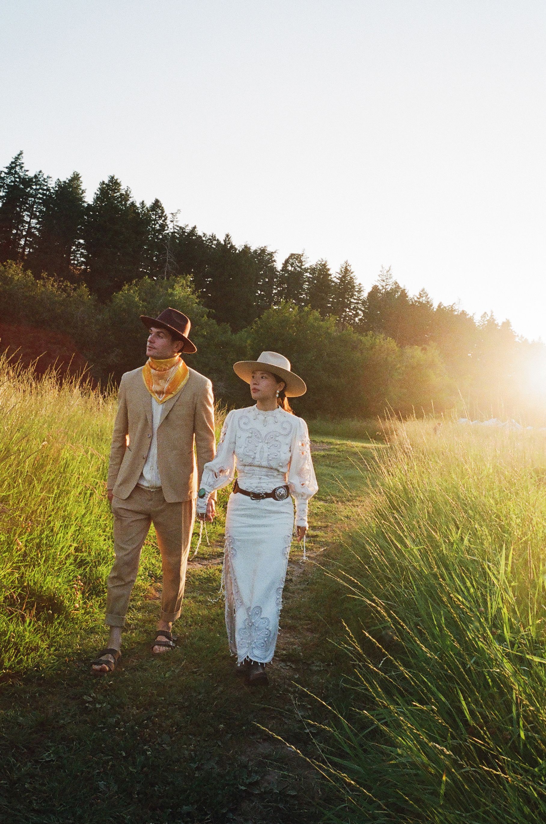 Two people walking through a sunlit grassy field near trees