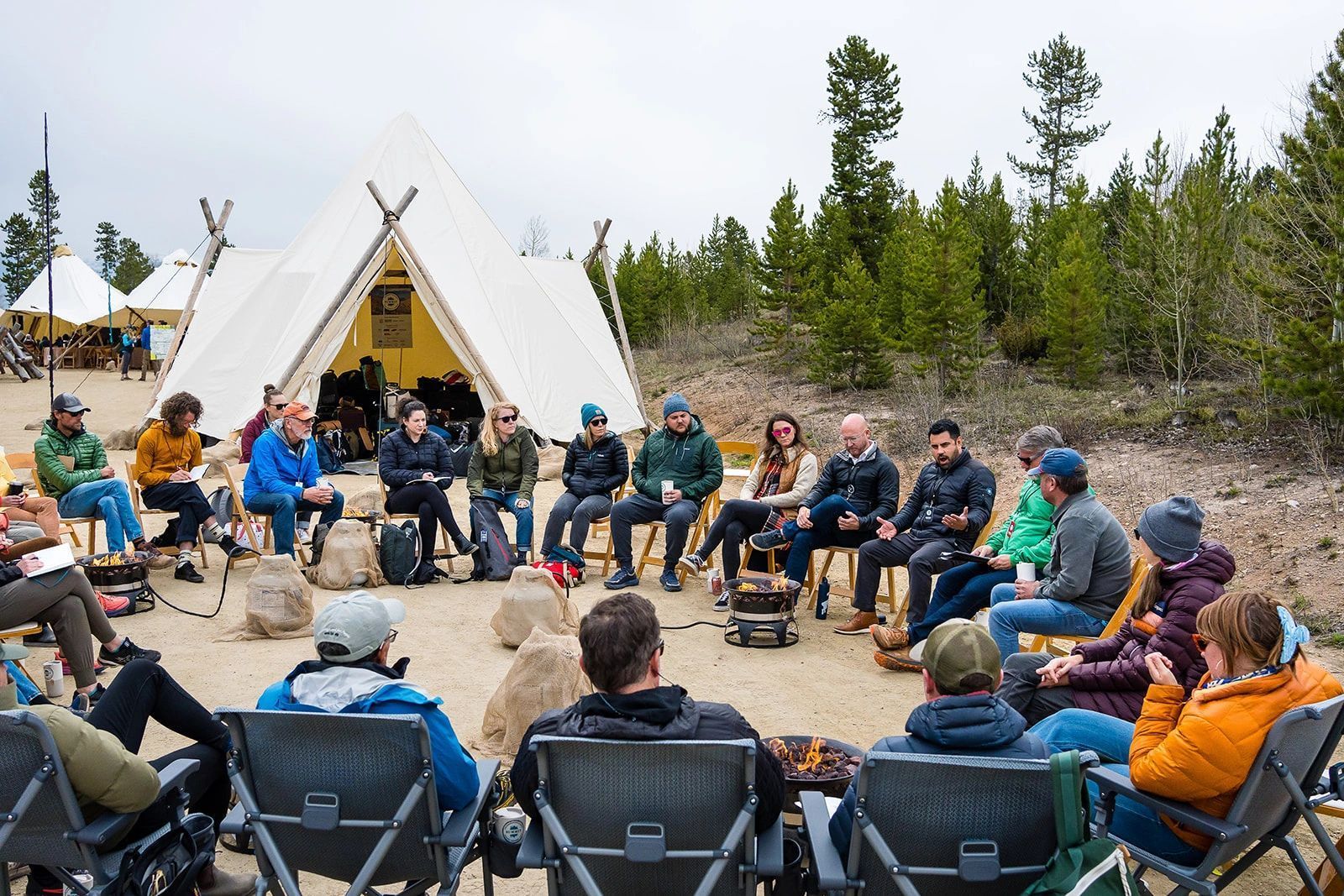 People seated in a circle outside a teepee, gathered for an outdoor group discussion in a forest clearing