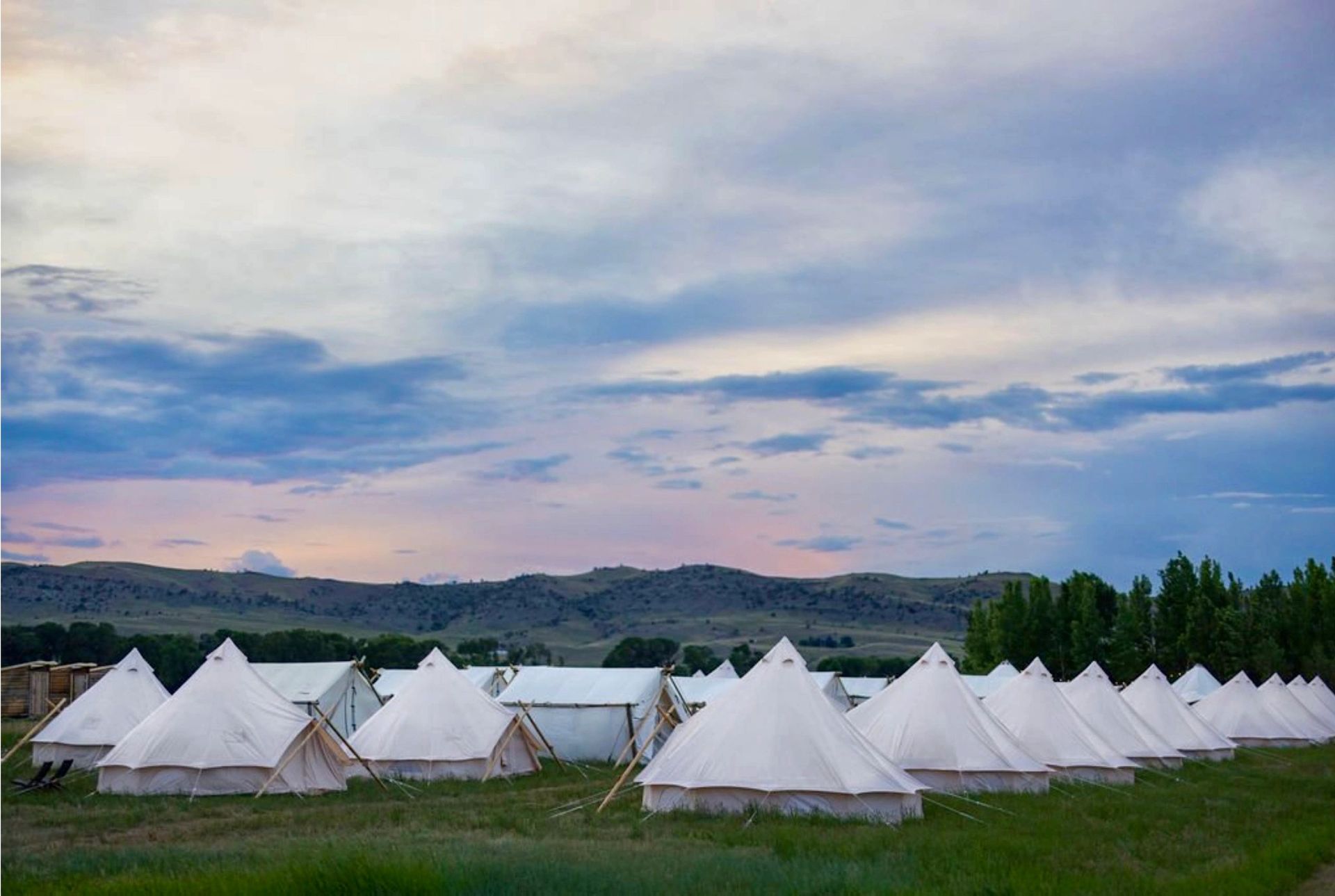 White teepees in a grassy field beneath a cloudy dusk sky with distant hills
