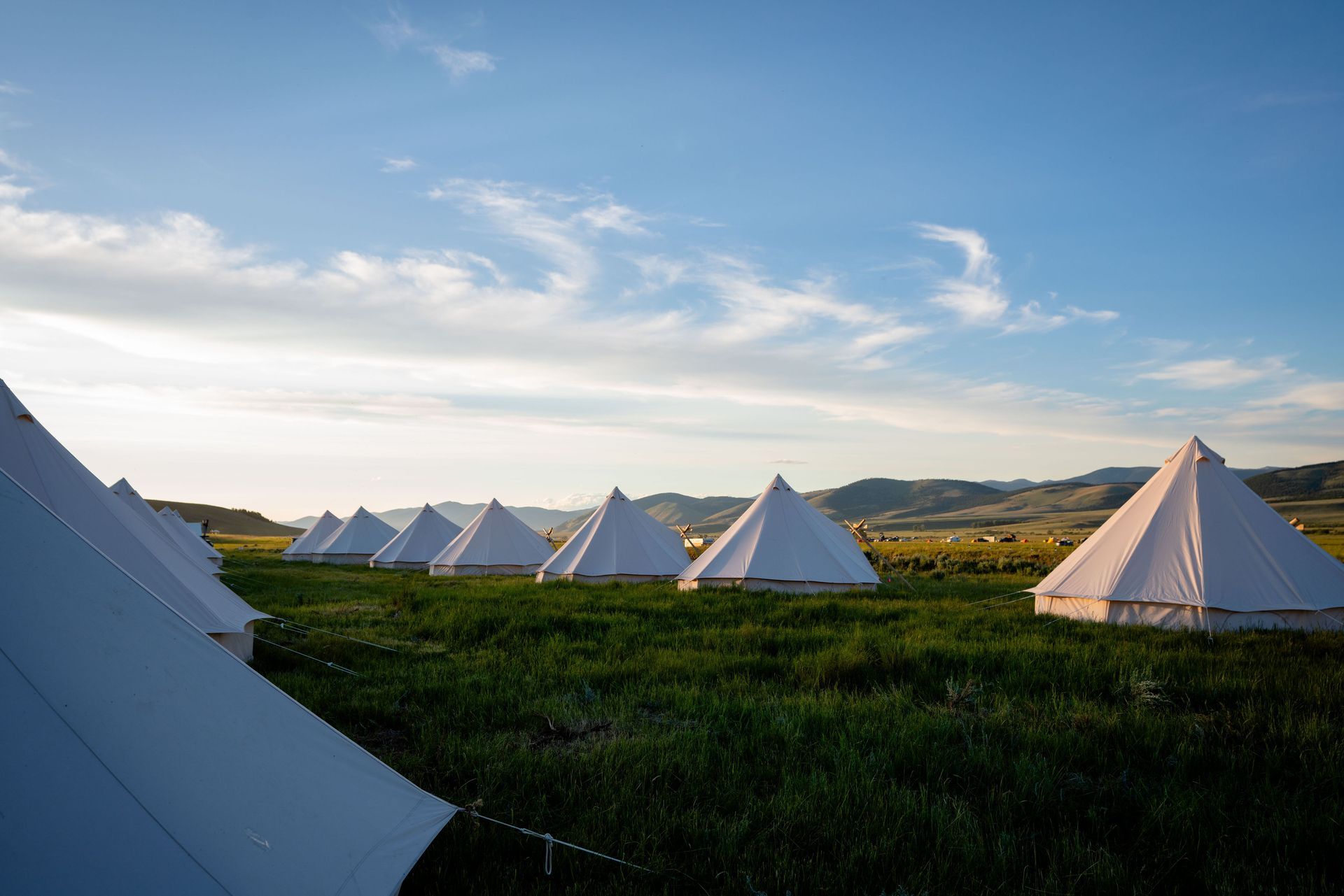 White teepee tents in a grassy field at sunset with mountains in the distance