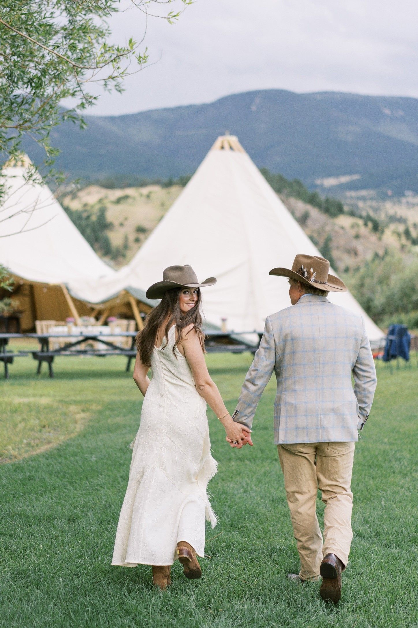 Couple holding hands, walking through a grassy outdoor wedding venue with white tents and mountains behind.