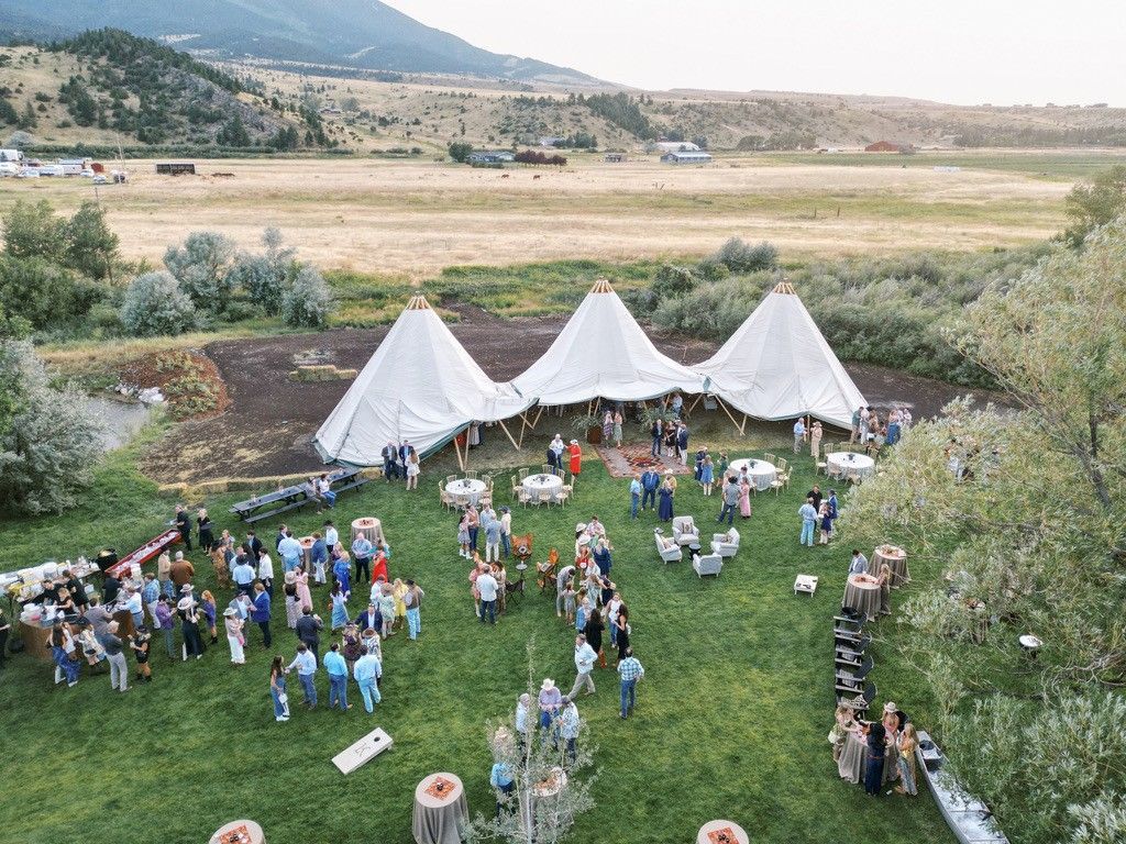 Outdoor gathering on a green lawn with white tents, tables, and many people, set against a mountain landscape