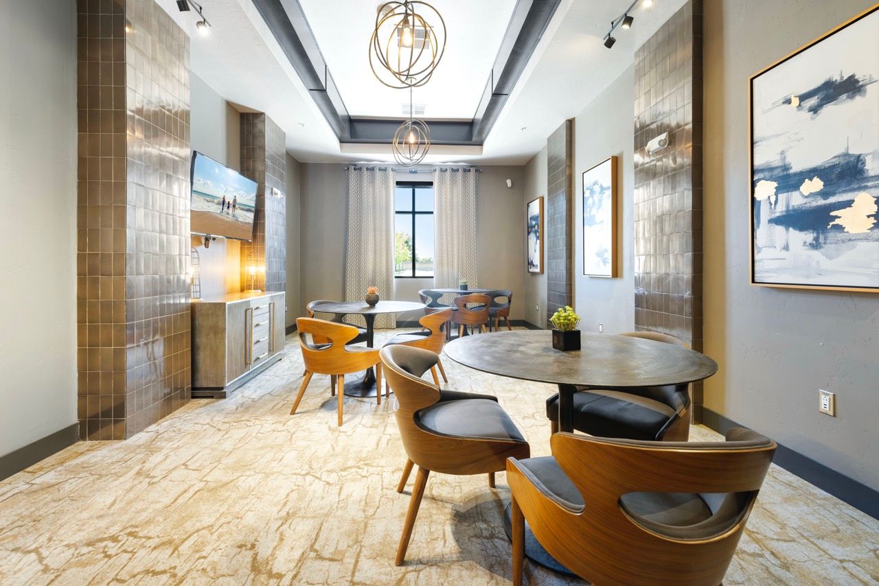 Indoor community lounge with round tables, wooden chairs, wall art, and a large window at Olympus Alameda, offering townhomes in Northeast Albuquerque, NM.