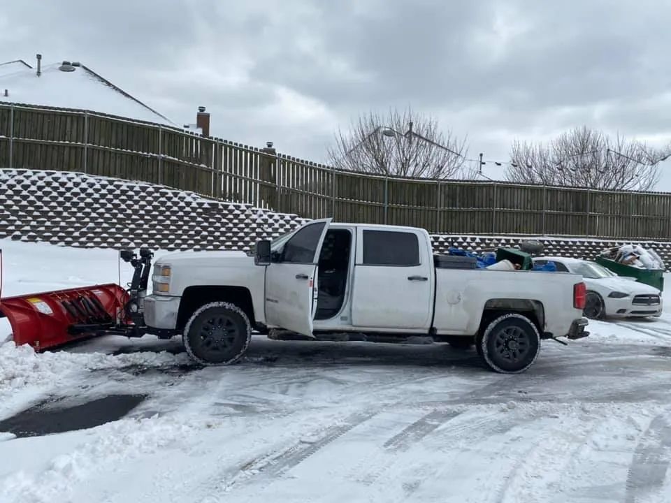 A white truck with a snow plow attached to it is parked in a snowy parking lot.