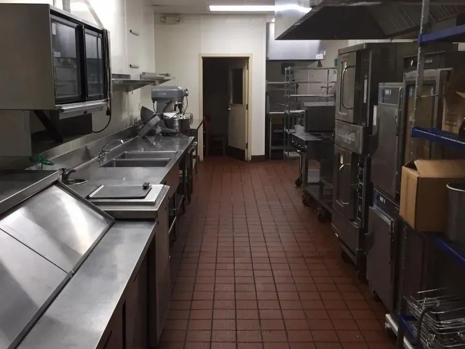 Empty commercial kitchen with stainless steel counters, ovens, and shelves along a tiled floor.