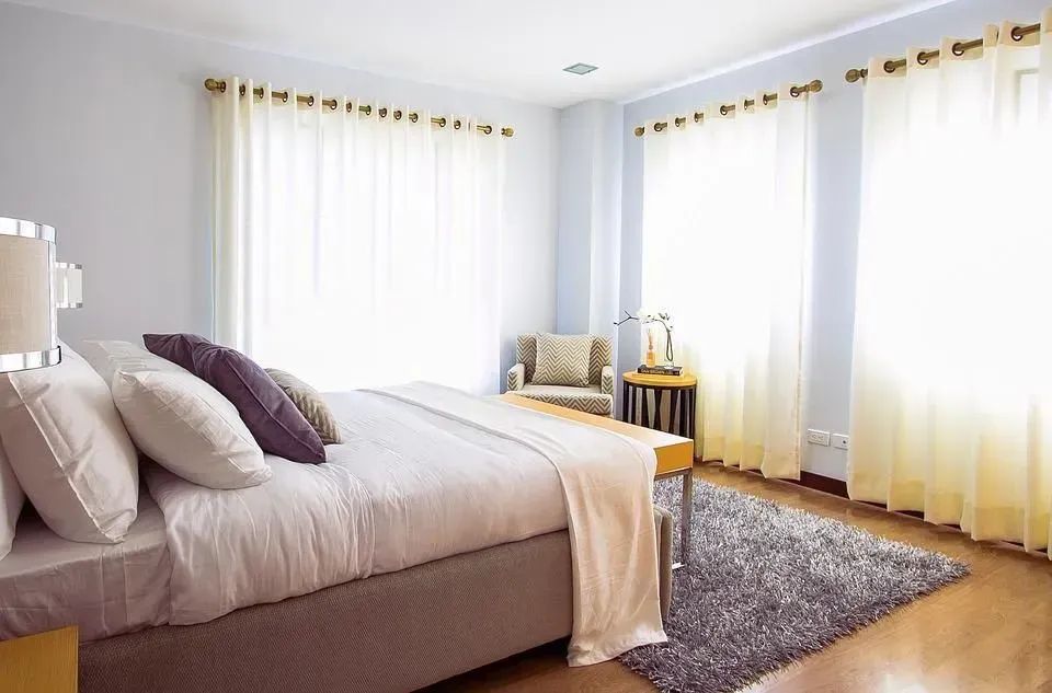 Bright bedroom with a neatly made bed, large windows, sheer curtains, and a gray rug.