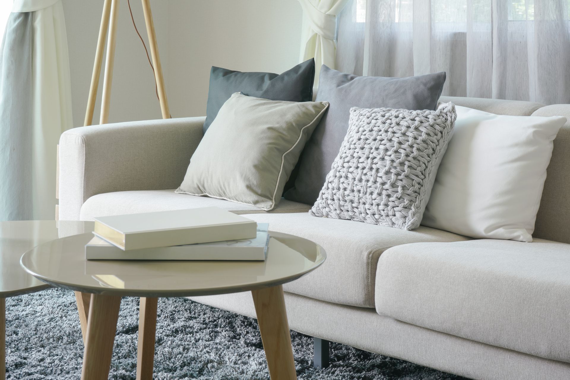 Beige sofa with pillows, round coffee table with books, window with sheer curtains.