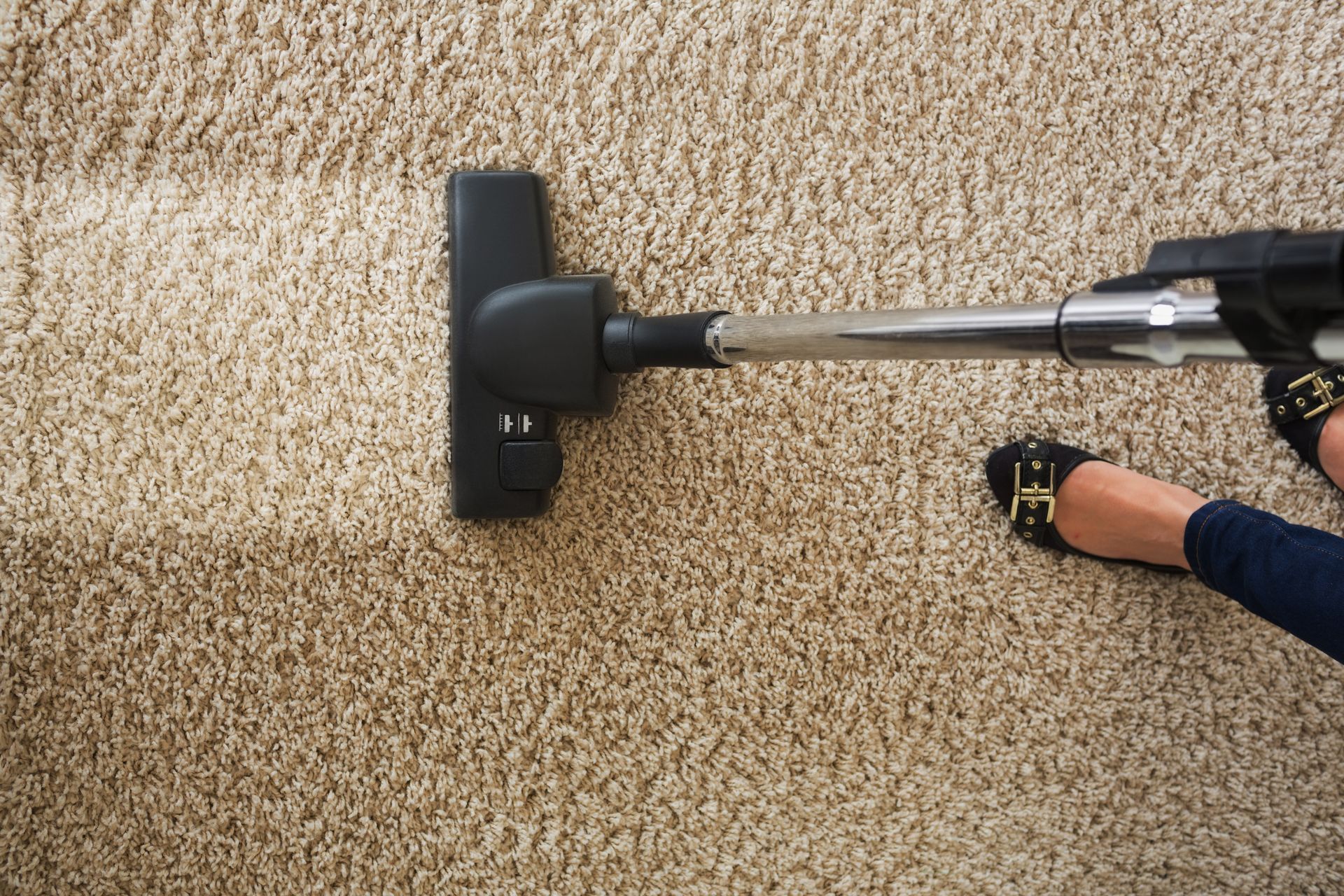 Vacuum cleaner on beige carpet beside a person’s foot and hand using the handle