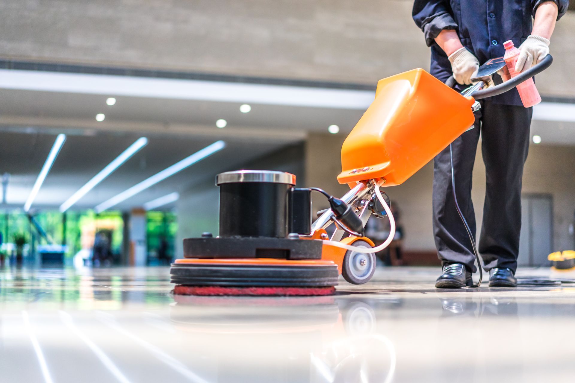 Person using an orange floor scrubber on a polished indoor floor in a modern building