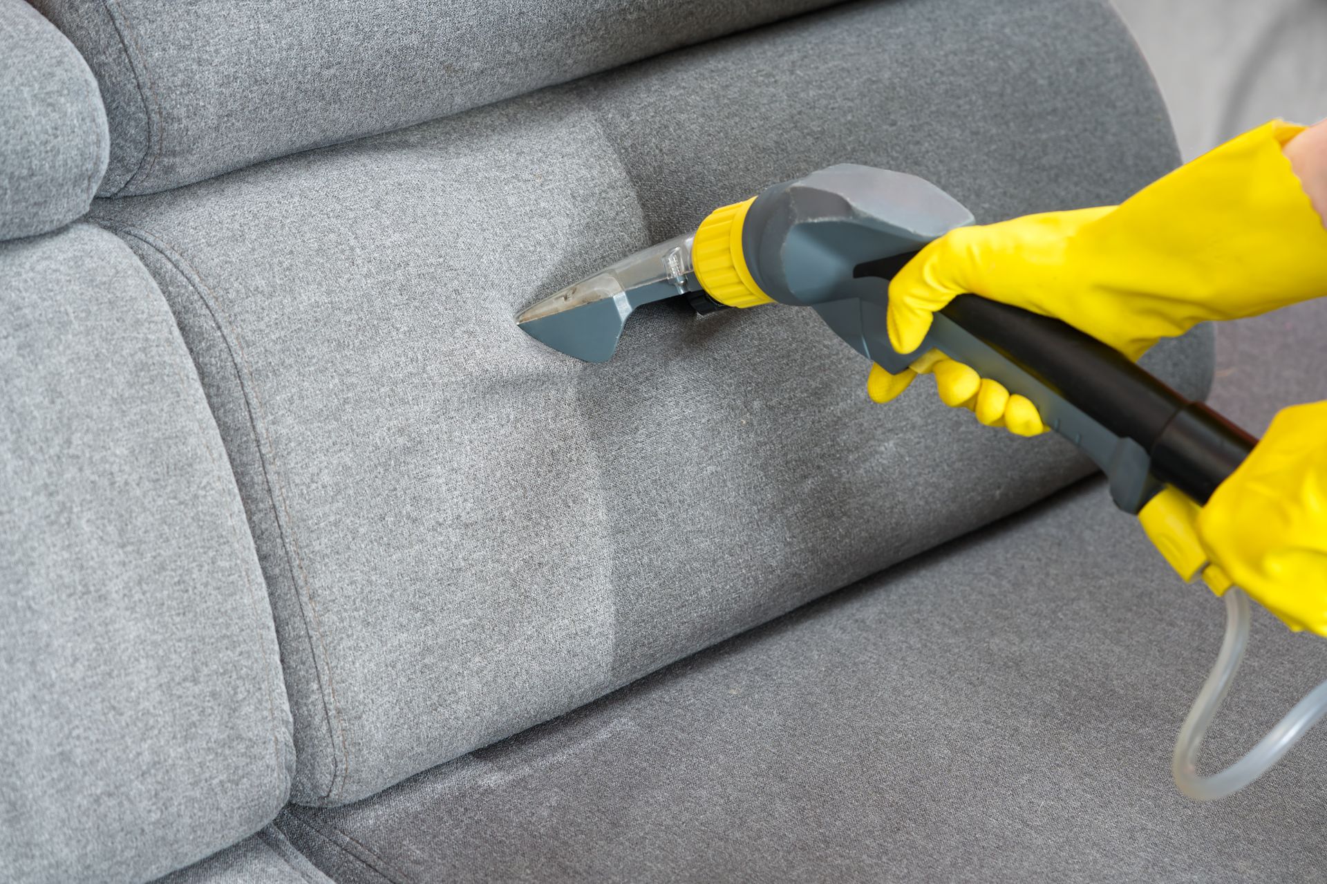 Yellow-gloved hand using steam cleaner on gray couch upholstery