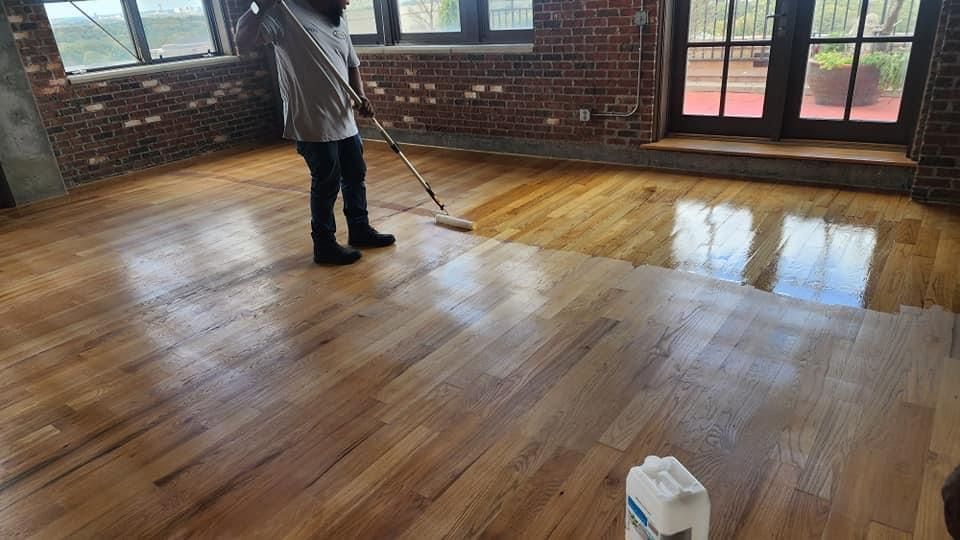 Person applying sealant to a hardwood floor with a roller. Shiny wood and exposed brick in a room.