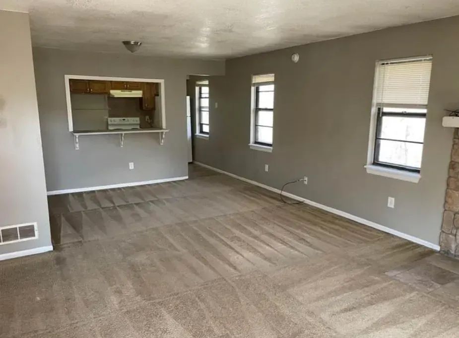 Living room with gray walls, tan carpet, windows, and an open kitchen view.