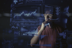 Person in glasses examining glowing financial charts on multiple screens in a dark room