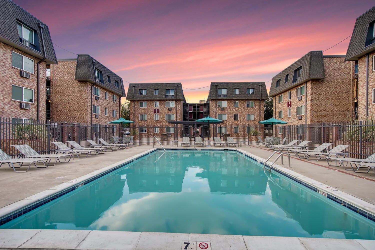Outdoor apartment complex pool with lounge chairs and fencing at sunset.