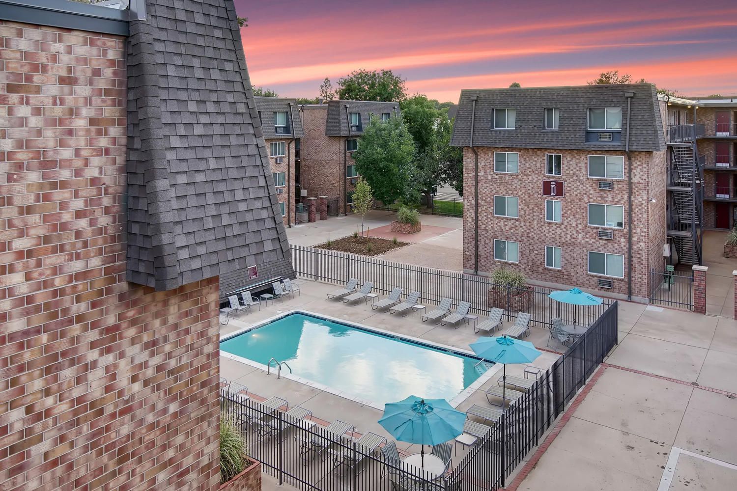 View of a communal apartment pool area with lounge chairs and blue umbrellas, surrounded by brick buildings.