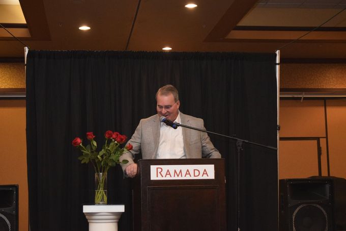 Man speaking at a podium with a microphone. He’s wearing a blazer, with red roses in a vase nearby.