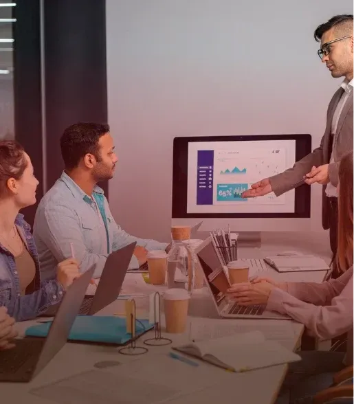 Business meeting with presenter pointing at a chart on a monitor as colleagues watch around a table