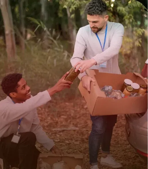 Two people exchange canned goods outdoors beside a car, with a box of food donations nearby.