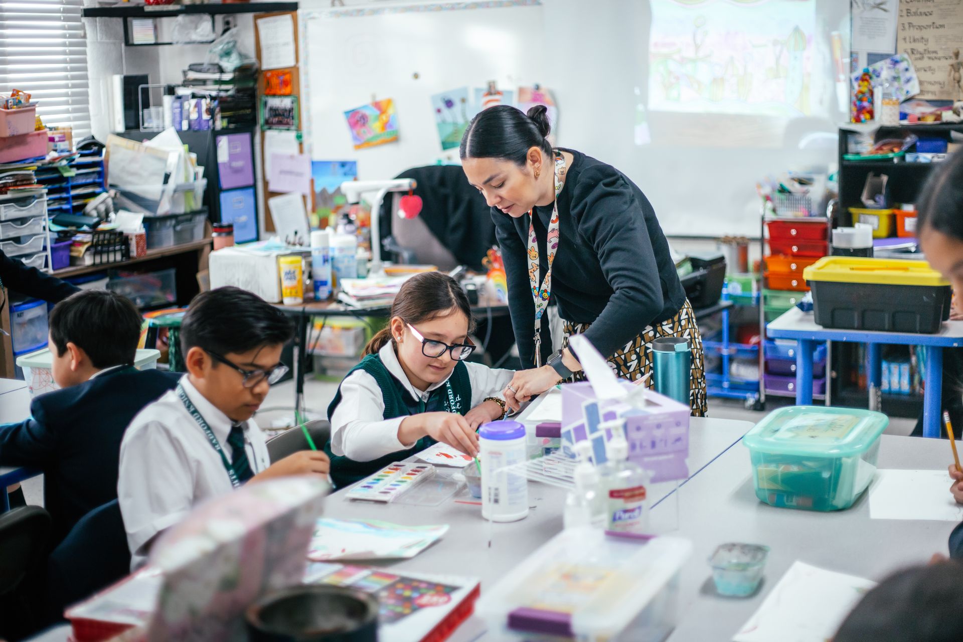 Teacher helping students in an art class. Students painting at tables, classroom setting.
