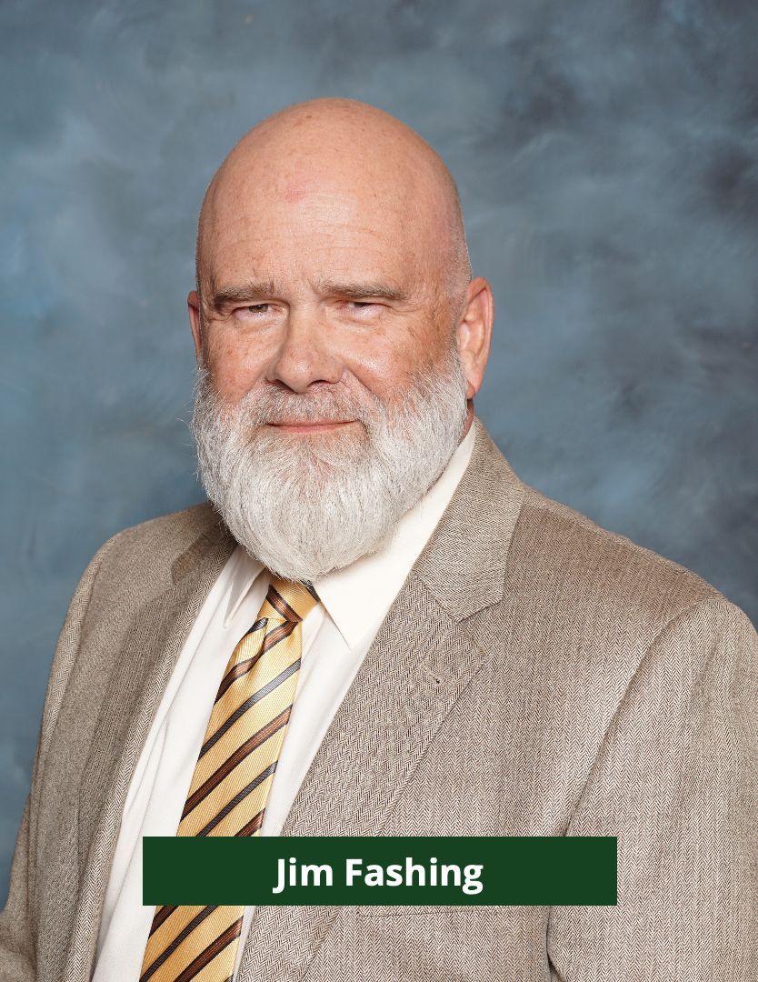 Bald man with white beard wearing a tan blazer, white shirt, and striped tie; green background.