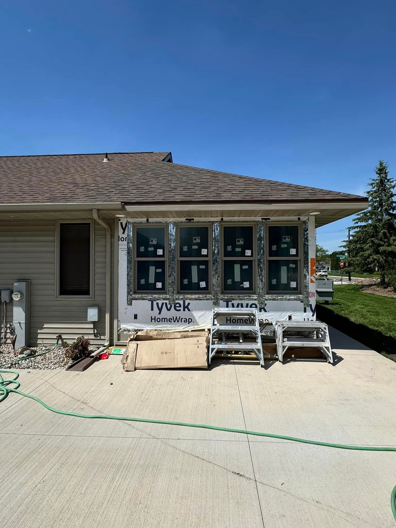House under construction with wrapped windows, roof shingles, and ladders on a concrete driveway under a blue sky
