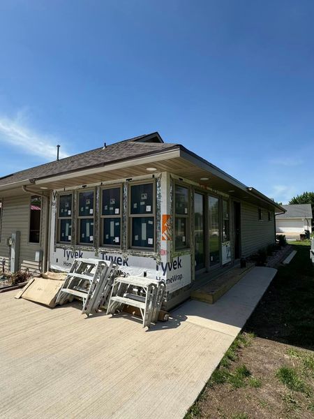 Small house exterior with large windows, white siding, and a porch with stacked chairs under a blue sky