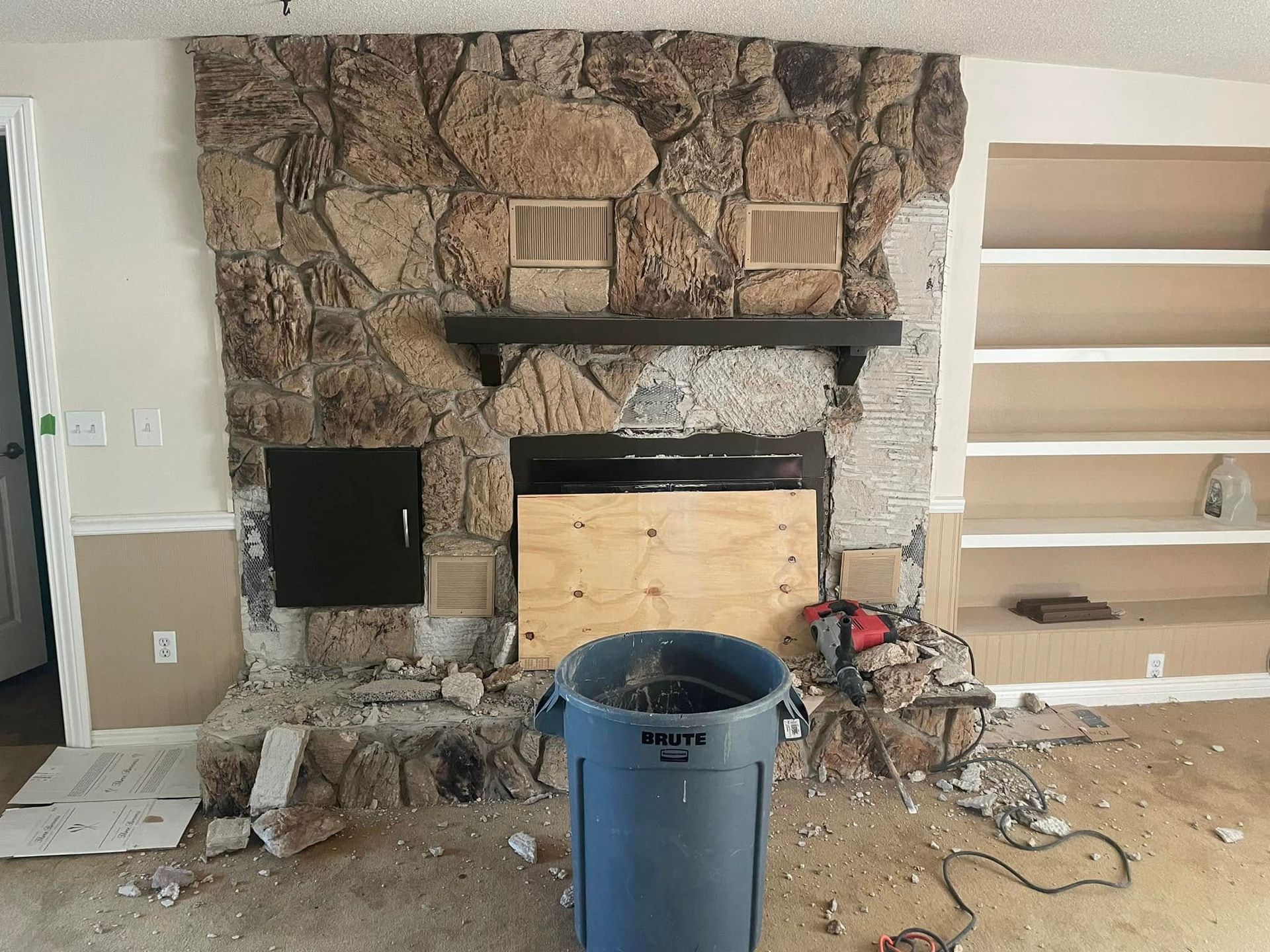 Living room with stone fireplace under renovation, debris on floor, blue trash can in foreground