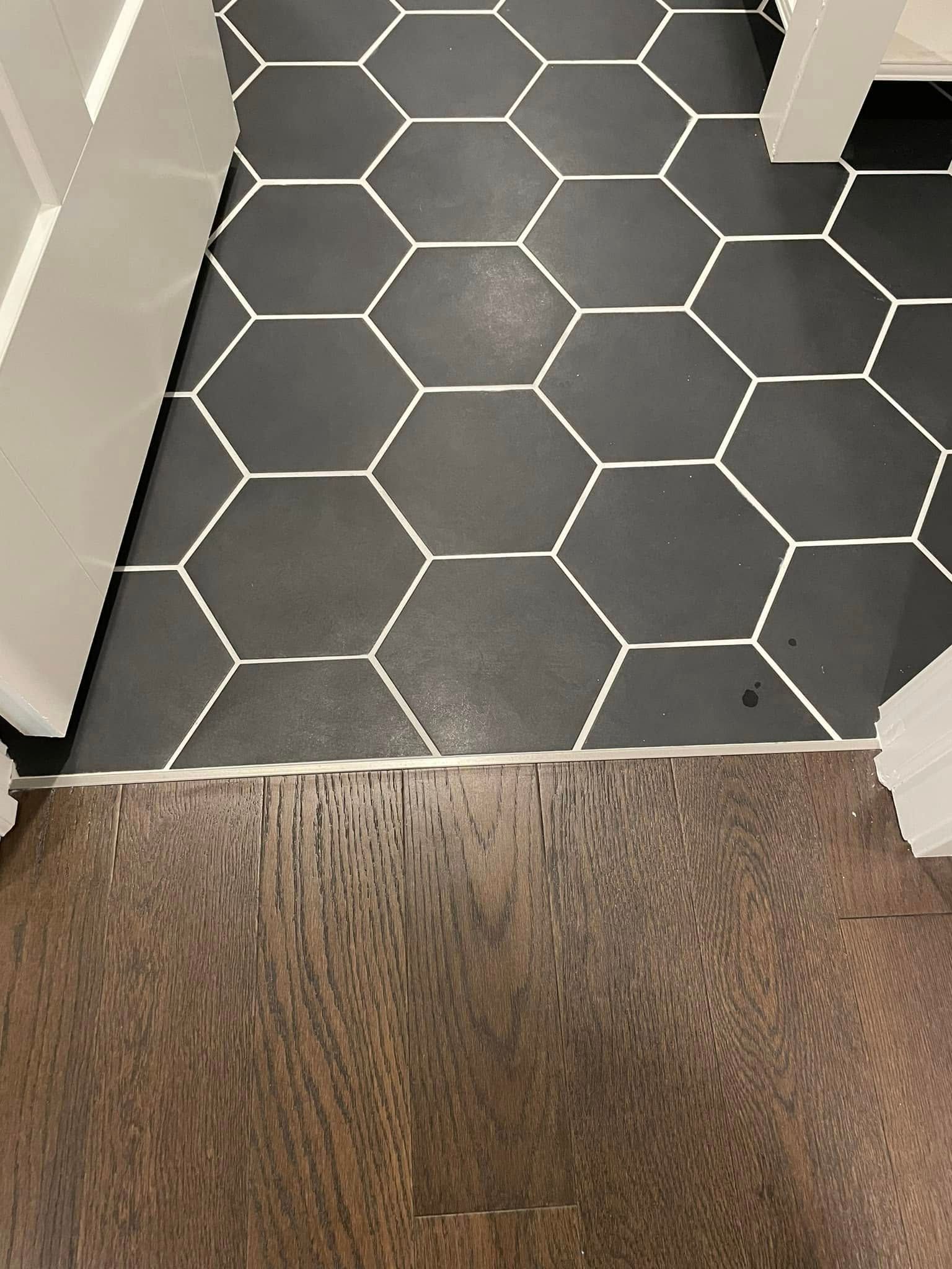 Black hex tile floor meets brown wood floor at a threshold beside a white appliance.