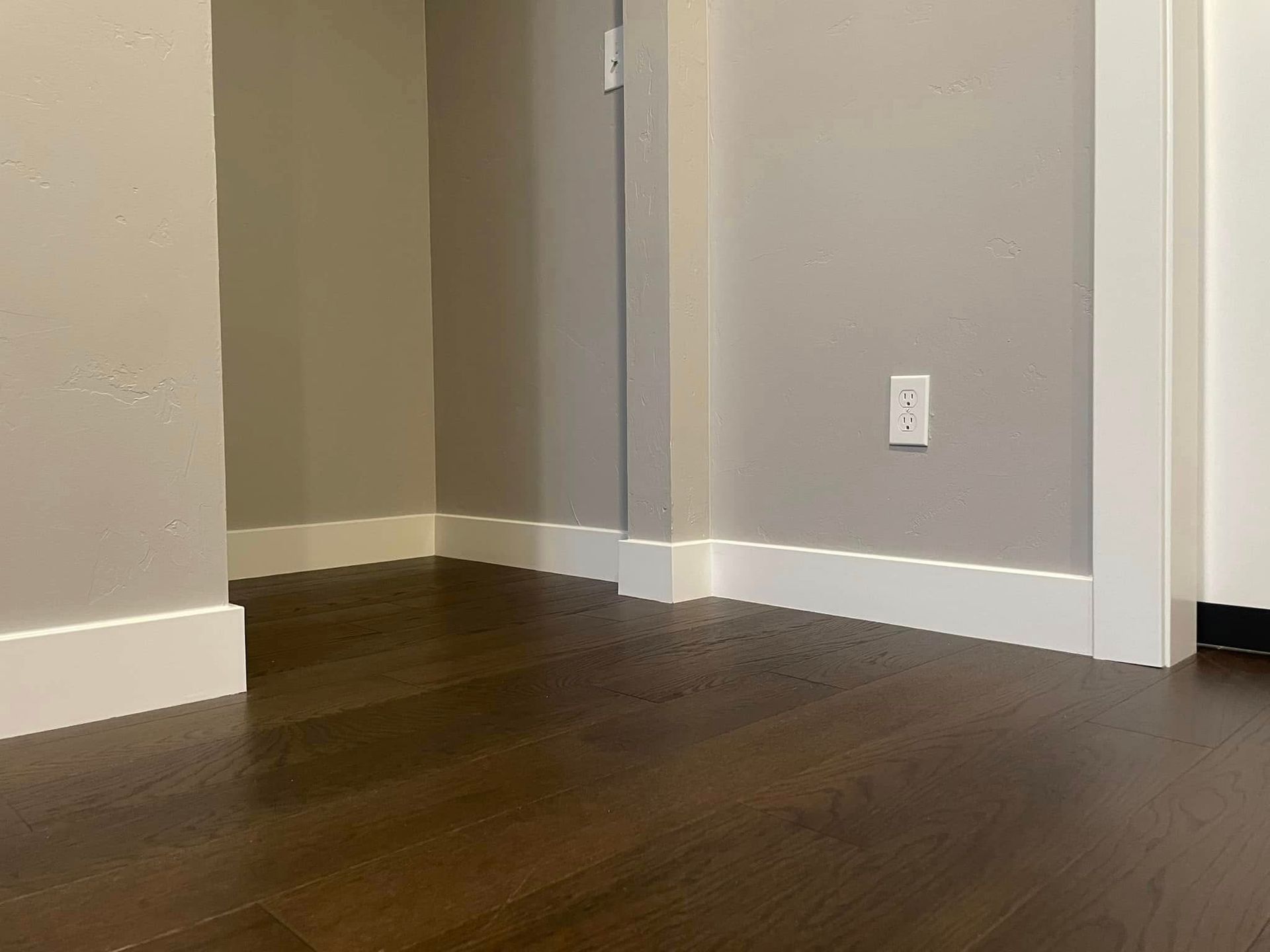 Empty room corner with dark hardwood floor, beige walls, white trim, and an electrical outlet.