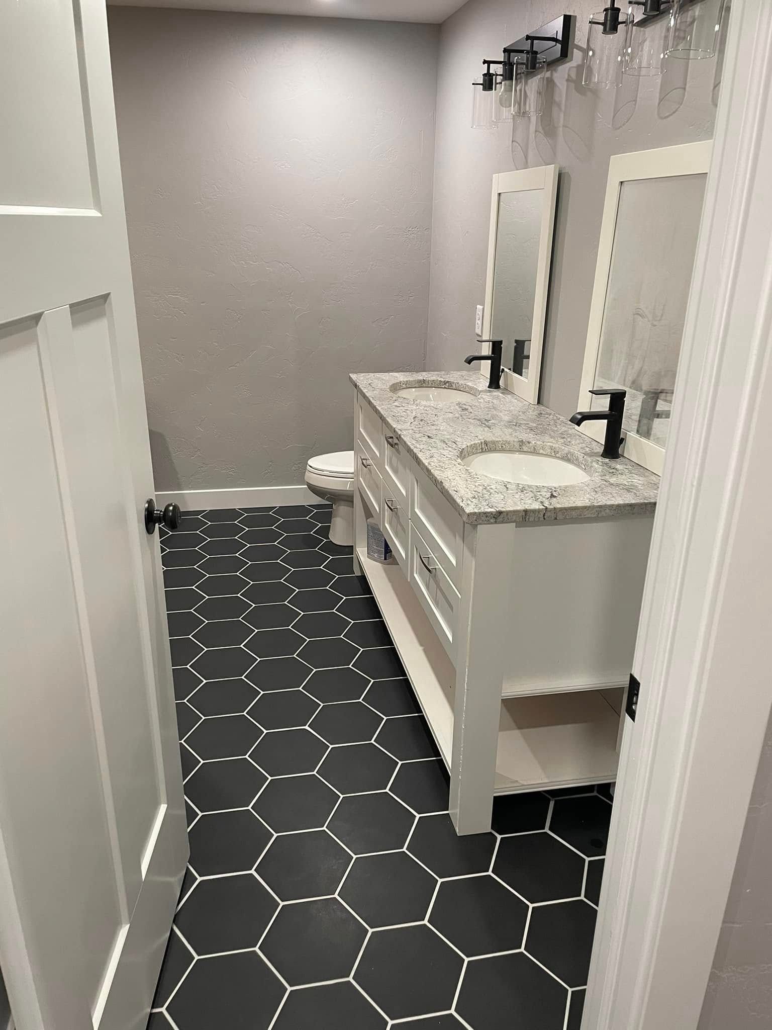 Small bathroom with black hex tile floor, white vanity, marble countertop, mirror, and toilet stall.
