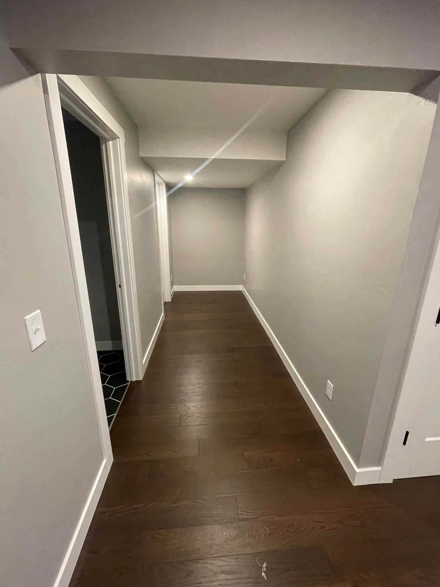 Empty hallway with gray walls, dark wood floor, and recessed ceiling lights leading to a doorway.
