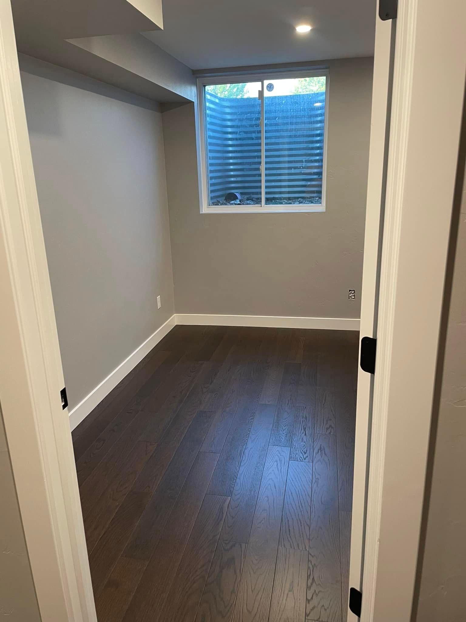 Empty narrow room with dark wood floor, gray walls, white trim, and a small window.