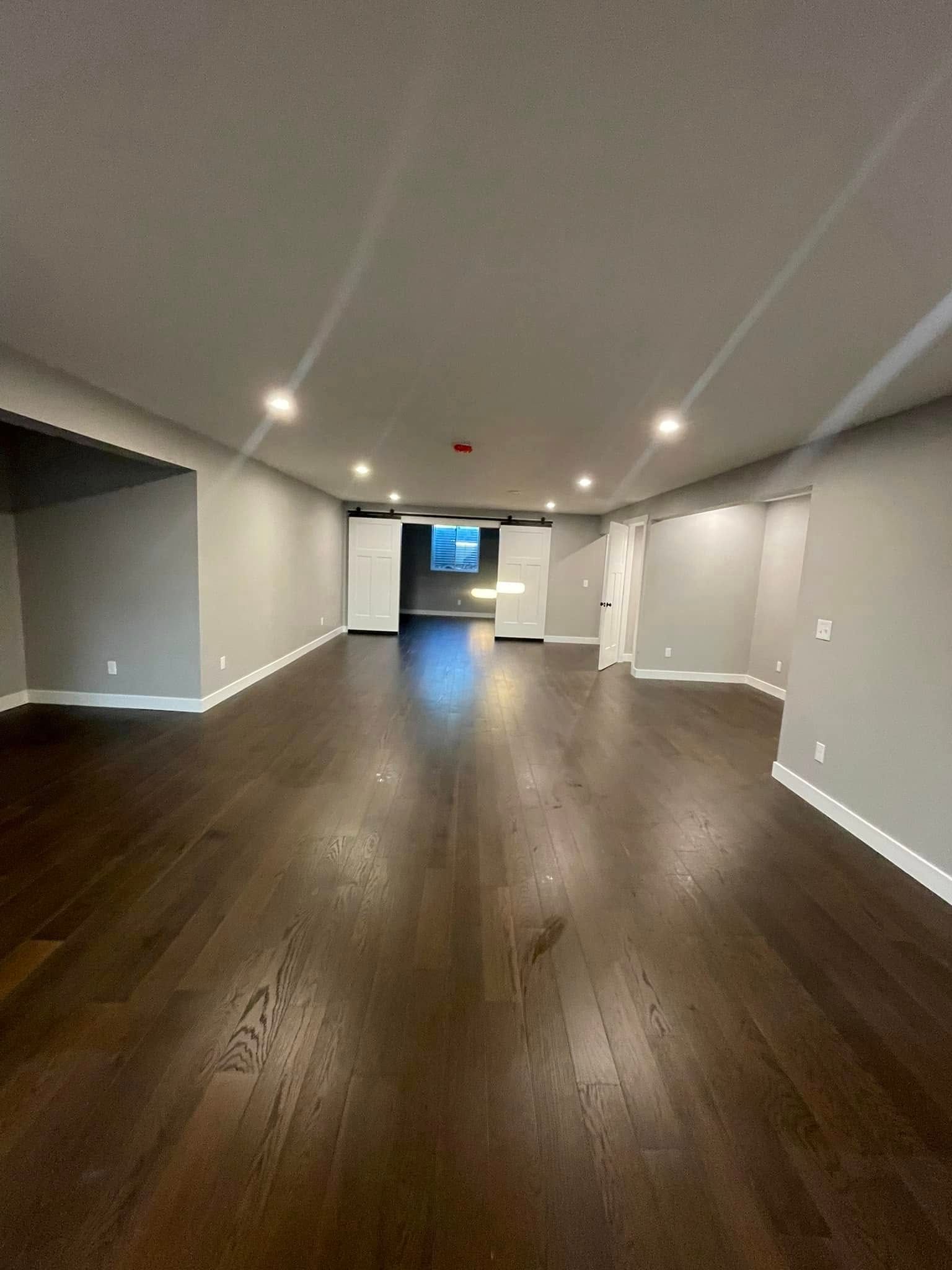 Empty modern living room with dark wood floors, gray walls, recessed lights, and a view into another room.