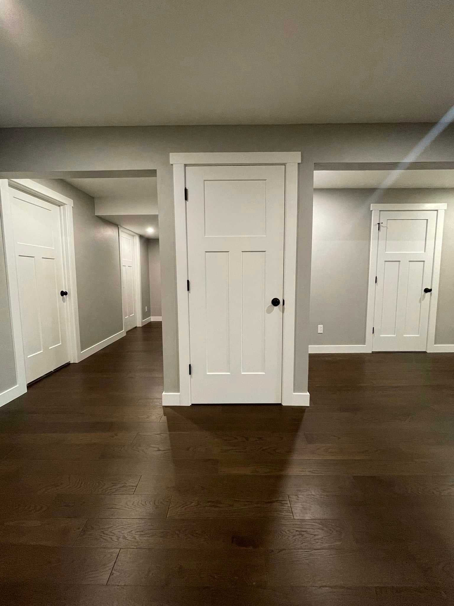 Empty carpeted room with dark wood floors, white walls, and several closed doors.