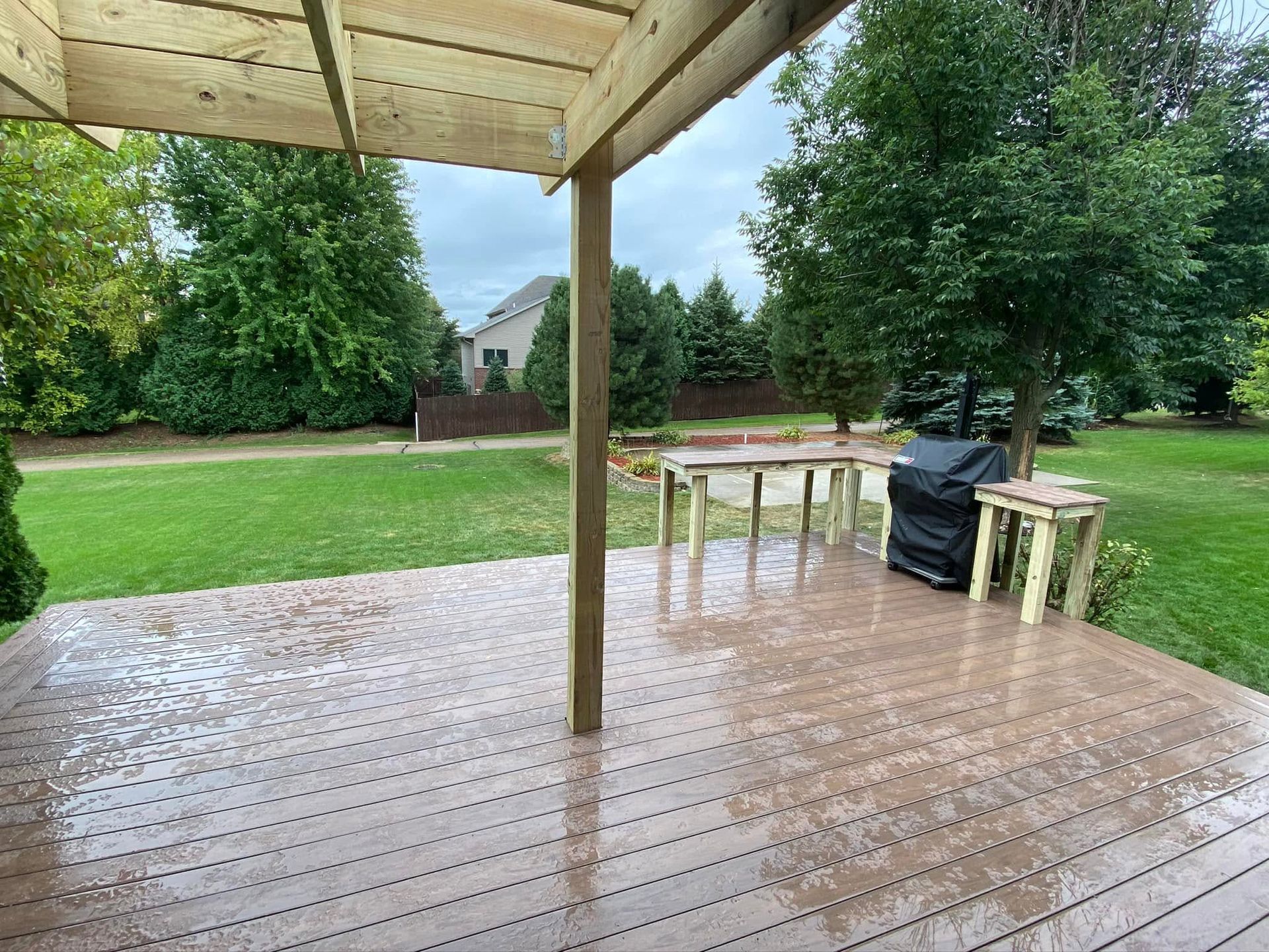Rainy backyard deck with wet wooden floor, patio table, grill, and grassy yard beyond