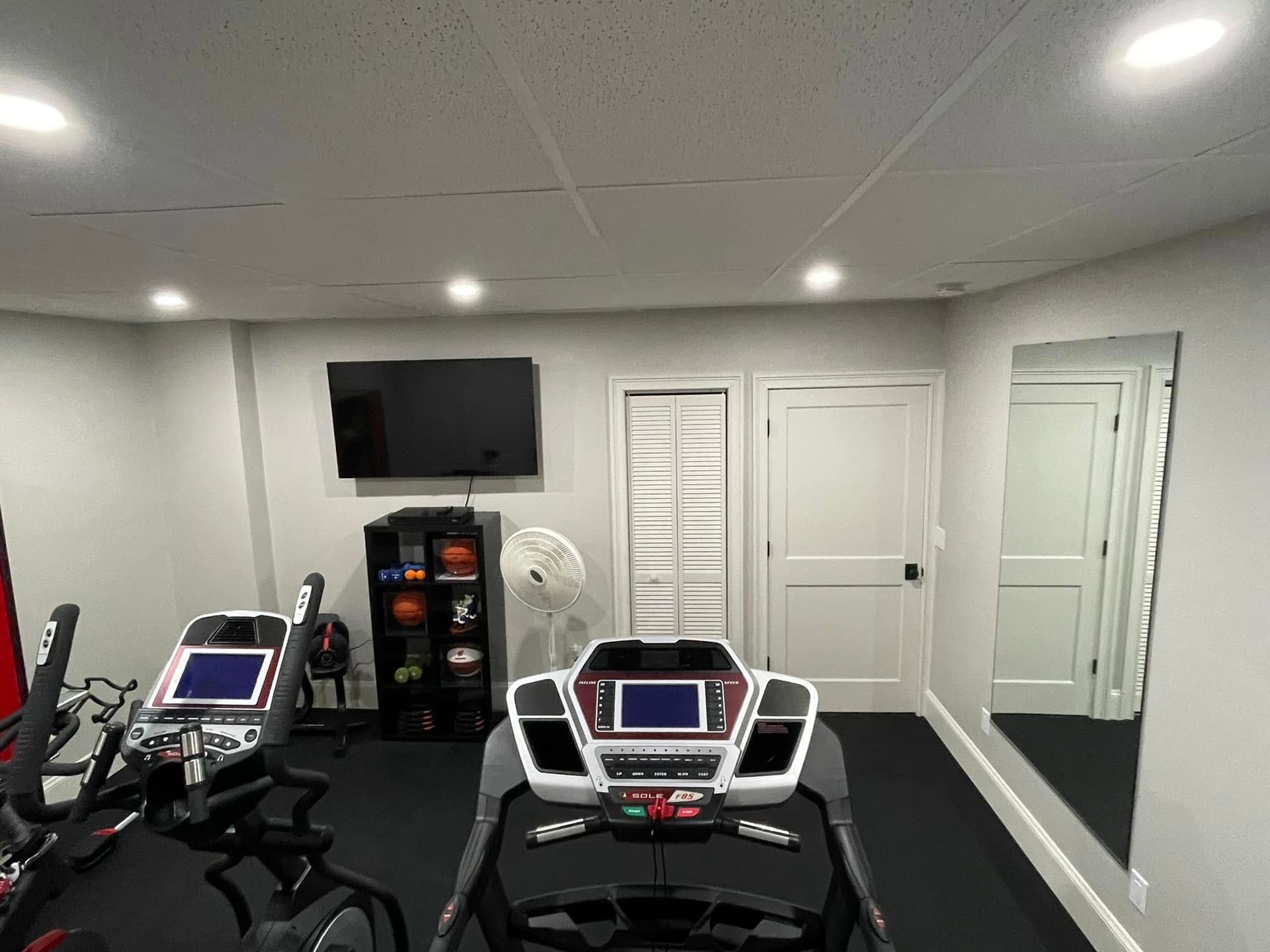 Gym room with two treadmills, mirrors, a fan, and white doors on the back wall.