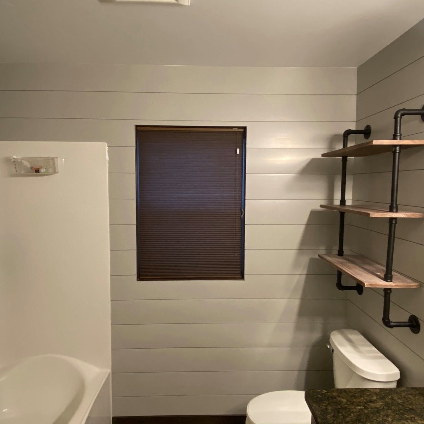 Small bathroom with white shiplap walls, dark window blind, tub, toilet, and black pipe shelving.