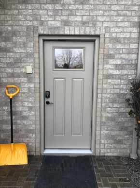 Gray front door in a brick wall, with a black doormat and a yellow snow shovel beside it.