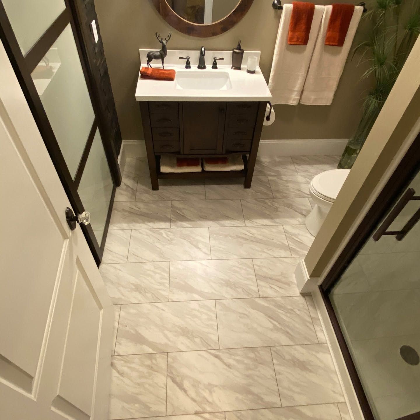 Small bathroom with a dark vanity, white sink, beige tile floor, and orange towels by the mirror.
