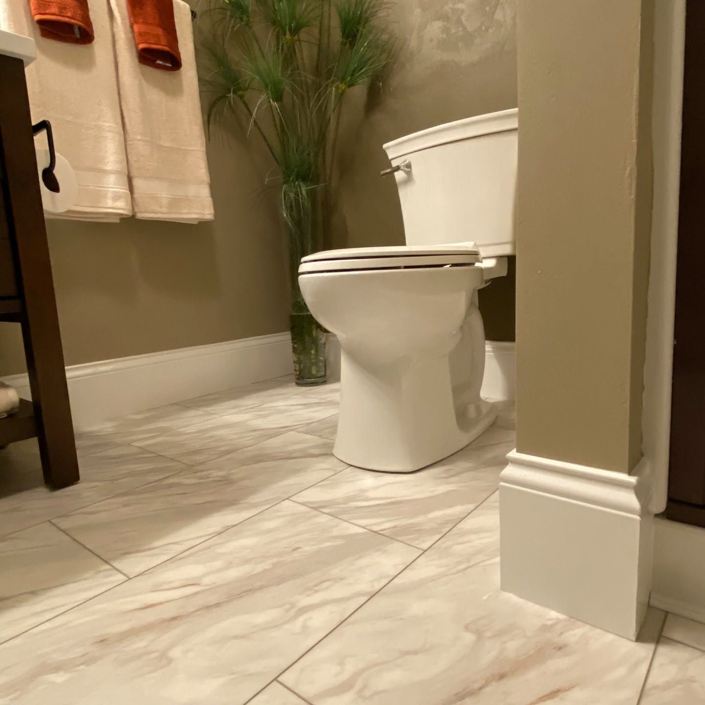 Bathroom with a white toilet, beige tile floor, towels, and a potted plant.