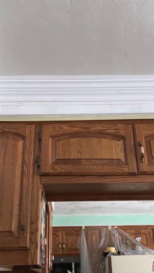 Wooden kitchen cabinets beneath white crown molding and a beige wall, with a doorway visible below.