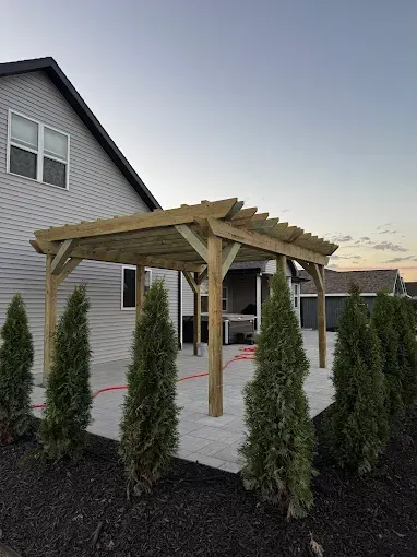 Wooden pergola entrance beside a house, framed by tall evergreen shrubs on a landscaped driveway.