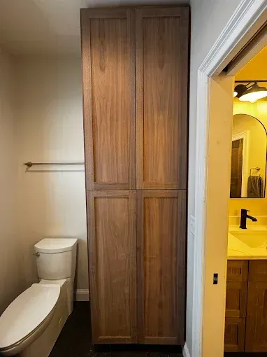 Small bathroom with toilet, tall wood cabinet, and open doorway to a yellow-lit sink area