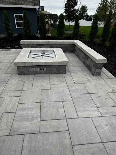 Modern gray stone patio with a square fire pit and built-in bench seating beside a house and lawn.