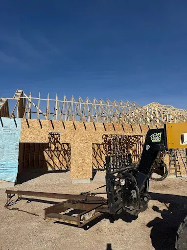 Wood-framed house under construction with exposed trusses and a forklift in the foreground