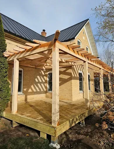 Wooden pergola deck attached to a brick house under construction, with a dark metal roof and snowy ground nearby