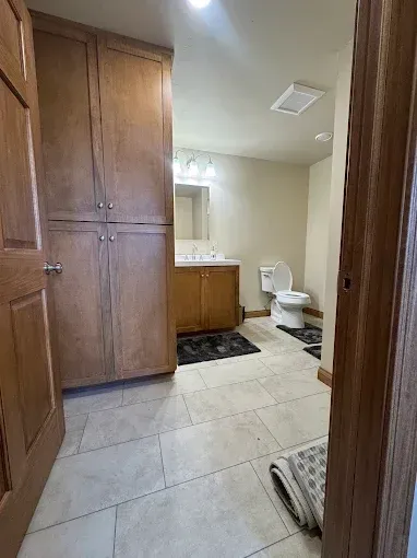 Small bathroom with wood vanity, toilet, white tile floor, and a rug by the doorway.