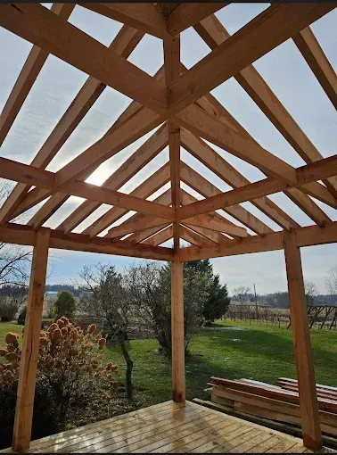 Wooden pergola frame with slatted roof over an open patio overlooking a grassy field and trees