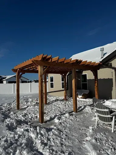 Wooden pergola in a snowy backyard beside a beige house under a clear blue sky.